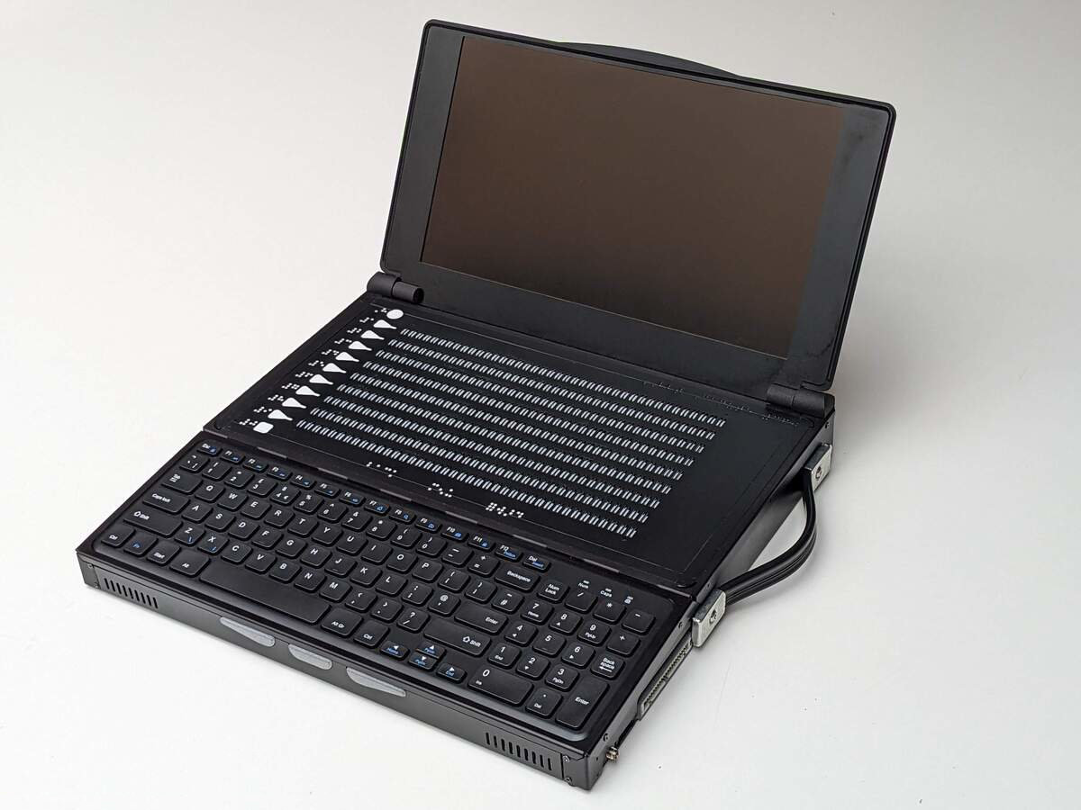Three quarters profile photo of the Canute Scientific when open. A full size keyboard in the front with a 360 cell Braille cell behind it. There is an open lid at the back with a 13" visual monitor. There is a heavy duty pull-out handle on the right side. The device is black with white details.
